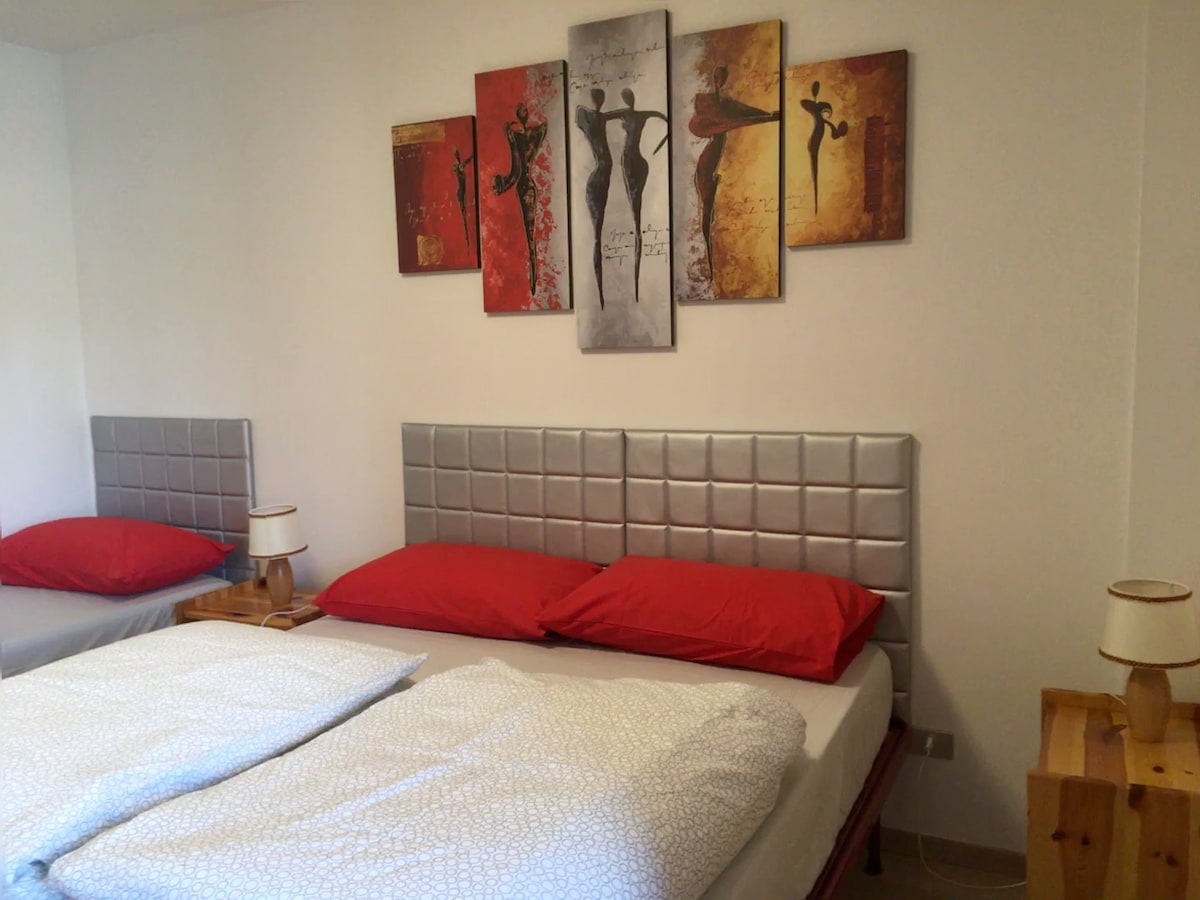 A spacious sleeping area features two beds with red pillows and a decorative wall artwork displayed above. Warm wood nightstands with lamps are positioned beside the beds, contributing to the overall cozy appearance of the room.