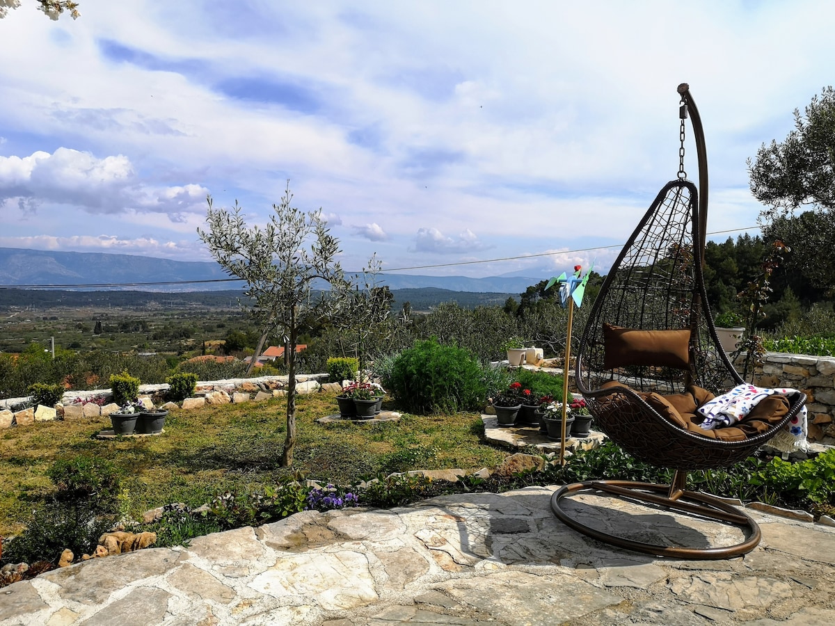 A hanging chair is positioned on a stone patio, providing a serene view of the surrounding landscape. Flower pots are arranged neatly throughout the garden, showcasing a variety of plants. The background features rolling hills under a partly cloudy sky.