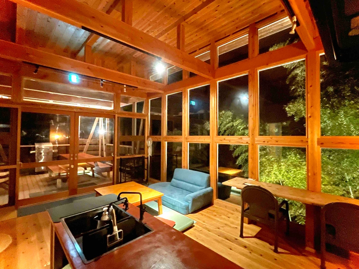 Wooden beams frame a spacious lounge area, illuminated by soft lighting. Large floor-to-ceiling windows provide views of the surrounding greenery, creating a connection with nature. A comfortable sofa and wooden tables invite relaxation in this tranquil environment.