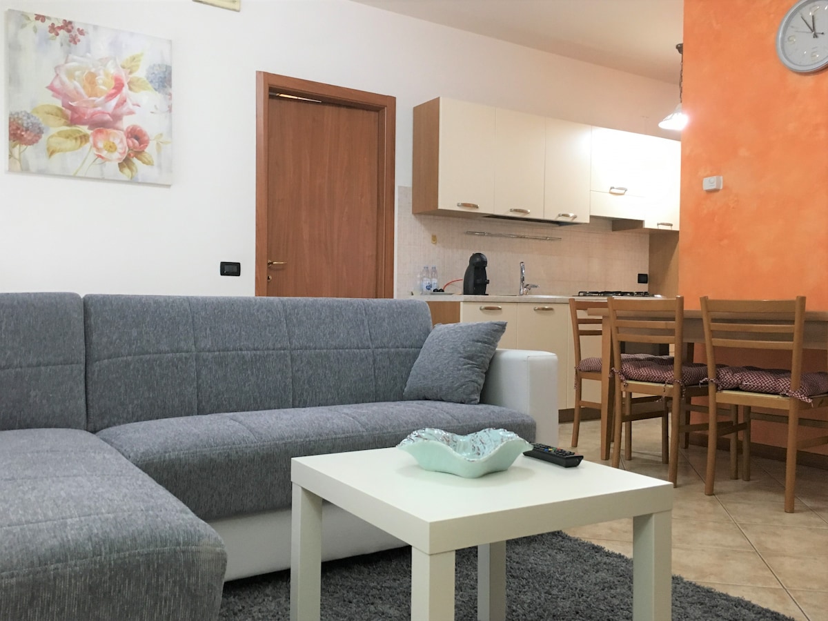 A bright living space features a gray sectional sofa positioned beside a small white table. A wall clock is mounted near an orange accent wall. The open kitchen, visible in the background, includes wooden cabinetry and a dining area with four chairs.