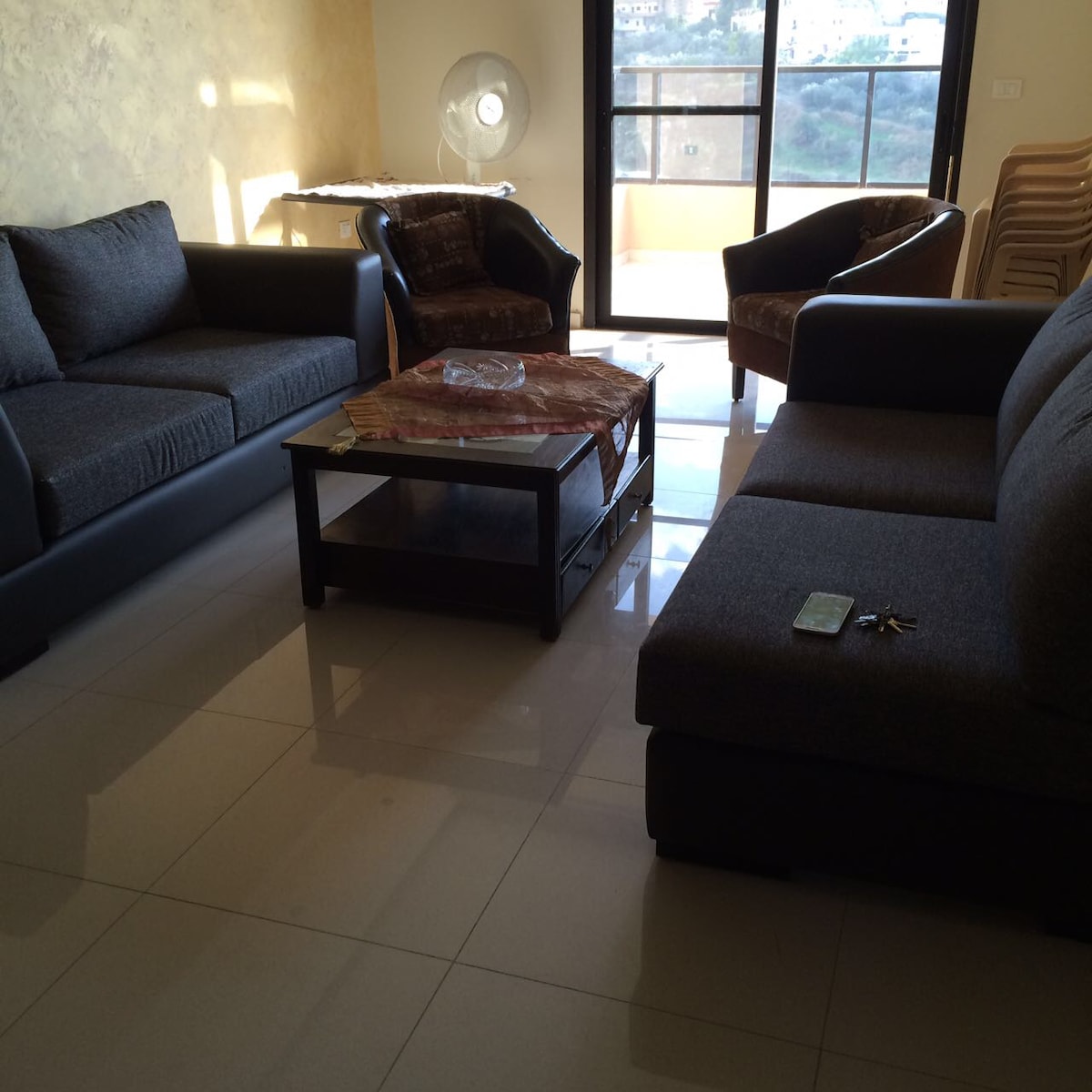 A living area is presented with a combination of modern seating, including a spacious grey sofa and two dark armchairs. A glass coffee table is centered on a reflective tile floor. Natural light filters through the large window, showcasing views of the surrounding landscape.