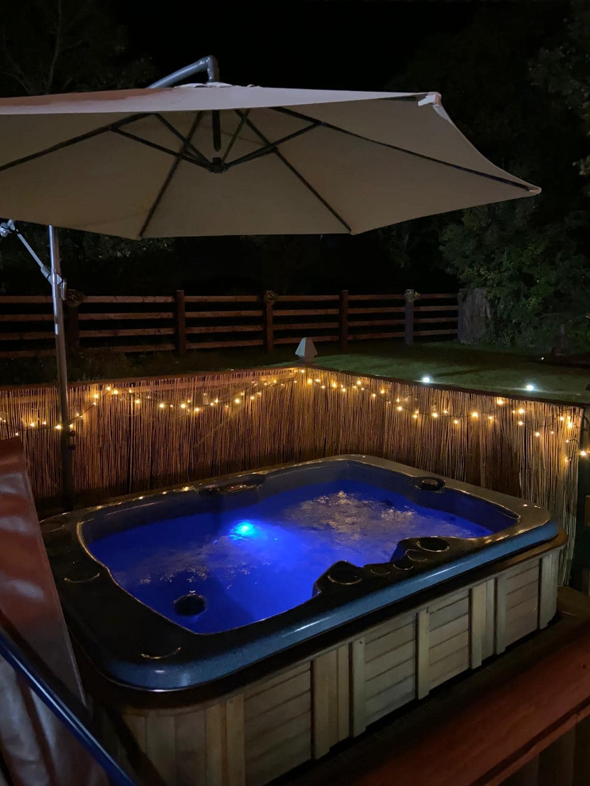 A relaxing hot tub is illuminated with soft blue lighting, surrounded by bamboo fencing. String lights add a cozy ambiance, while an umbrella provides shade. The secluded outdoor area is set against a backdrop of lush greenery, creating a tranquil space for guests.