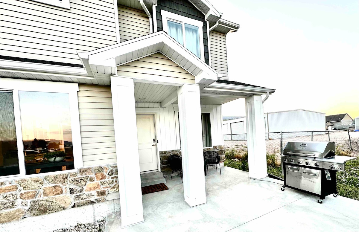 The exterior of the townhome features a welcoming entry with a covered porch, framed by two white columns. A gas grill is positioned on the patio, with a spacious driveway and landscaped area visible in the foreground, providing ample outdoor space.