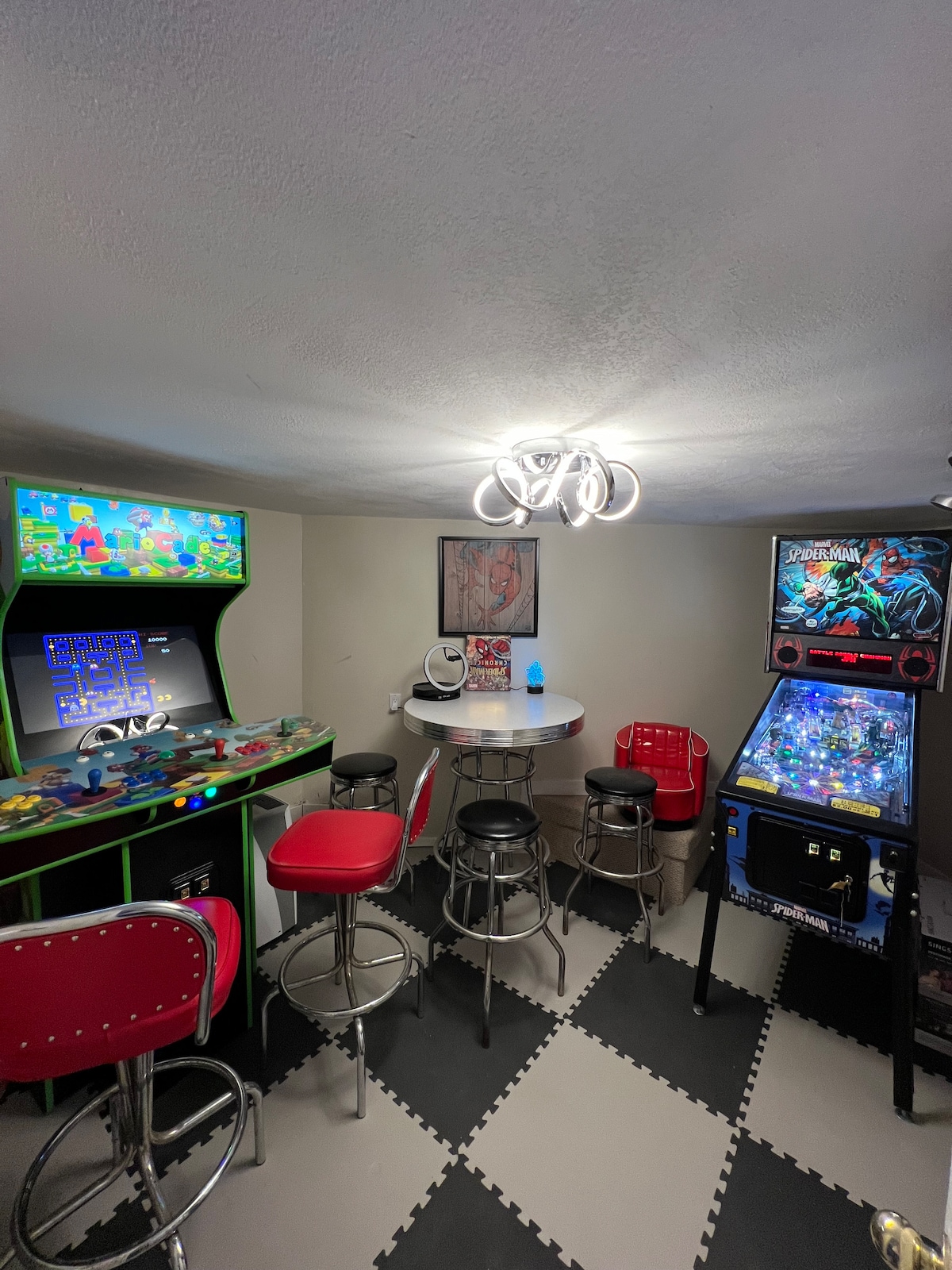 An arcade space is shown, featuring a colorful arcade machine and a pinball machine. Two red barstools are situated around a small table illuminated by a modern light fixture. The floor is covered in a black and white checkerboard pattern.