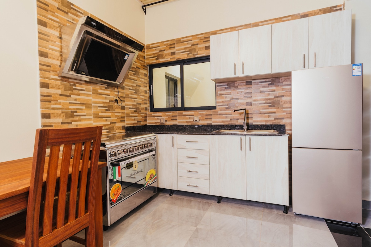 The kitchen features modern cabinetry with a sleek finish, complemented by a stone-patterned backsplash. A stainless steel refrigerator and an oven with a stovetop are visible. A wooden dining table with matching chairs is situated nearby, providing a functional dining space.