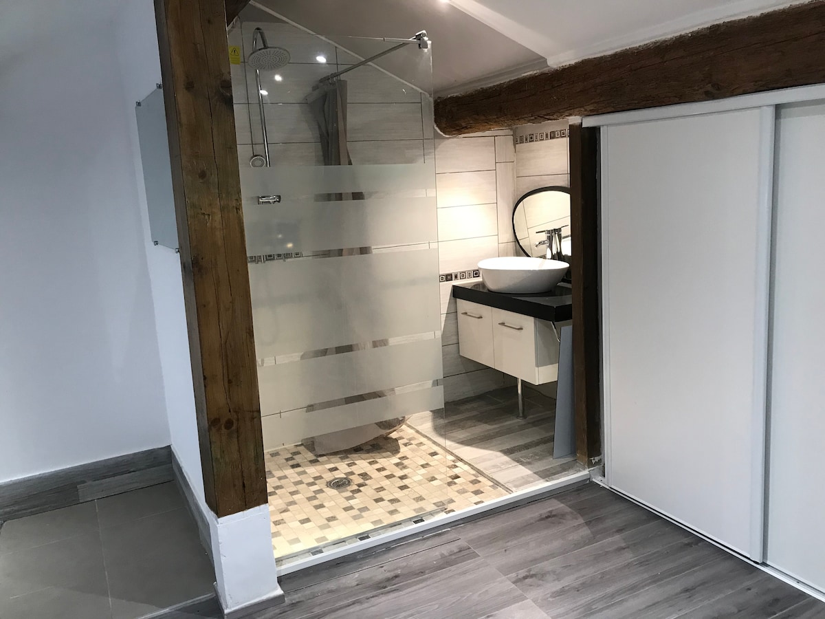 A modern bathroom is featured, showcasing a walk-in shower with frosted glass doors. A white sink is mounted on a dark vanity below a round mirror. Natural light is provided by overhead lighting and the wooden beams add a rustic touch to the design.