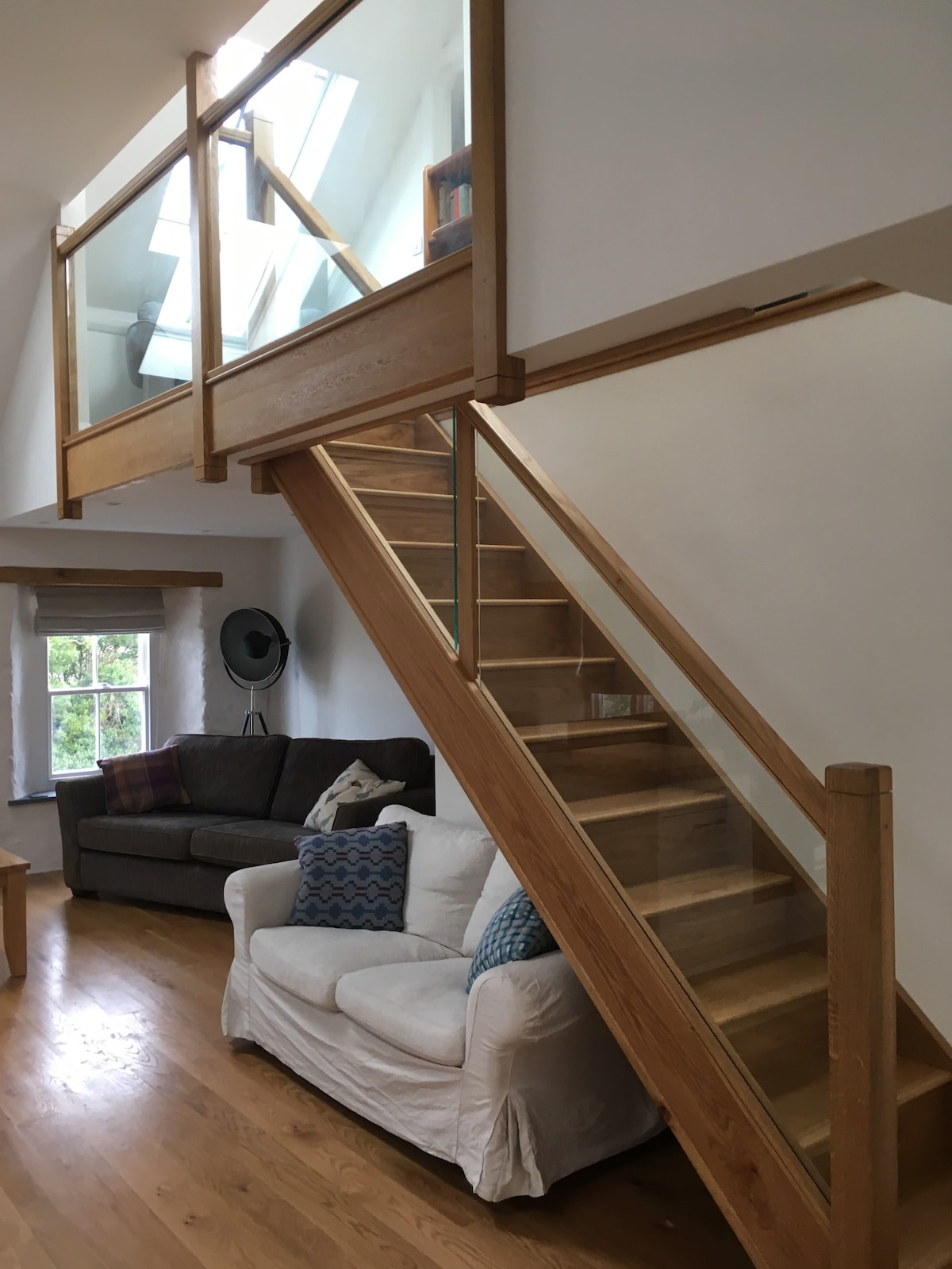 An open staircase made of oak leads to a loft area, featuring a glass railing that enhances visibility. Below, a cozy sitting area includes a dark brown couch and a light-colored, slipcovered chair adorned with blue patterned cushions. Natural light filters in from nearby windows.