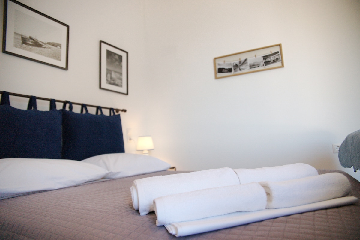 A spacious bed is positioned against a white wall, adorned with dark blue pillows. Neatly folded white towels are placed at the foot of the bed. Black and white photographs hang on the wall, complemented by a small lamp providing soft lighting.
