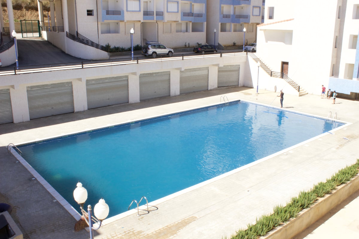 A large swimming pool is visible, surrounded by a spacious deck equipped with lounge areas and lighting fixtures. Umbrellas and potted plants line the edges, creating an inviting space. In the background, a multi-story residential building stands, with parking spaces and additional amenities also visible.