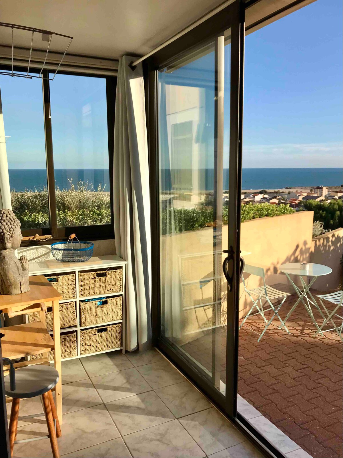 A sunlit veranda offers a panoramic view of the sea through large glass doors. Light-colored tiled flooring is complemented by a small table and chairs for outdoor dining. Storage baskets add a touch of functionality, while blue accents are present, enhancing the room's serene atmosphere.