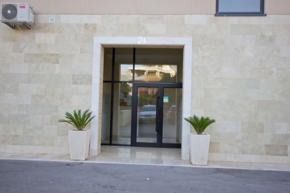 The building entrance is framed by two white planters containing small, green plants. A large glass door allows visibility into the lobby area, while the exterior features smooth stone walls and a clear contrast of texture. An air conditioning unit is visible on the left.