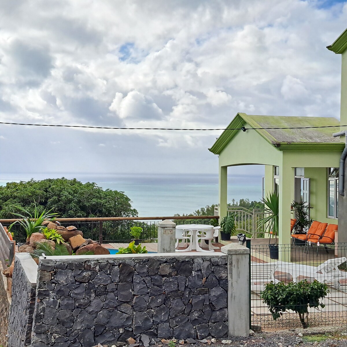 The image displays a charming green guesthouse with a white outdoor table and orange chairs positioned near a landscaped garden. In the background, the serene sea view meets a cloudy sky, while a private pool is visible beside the house.