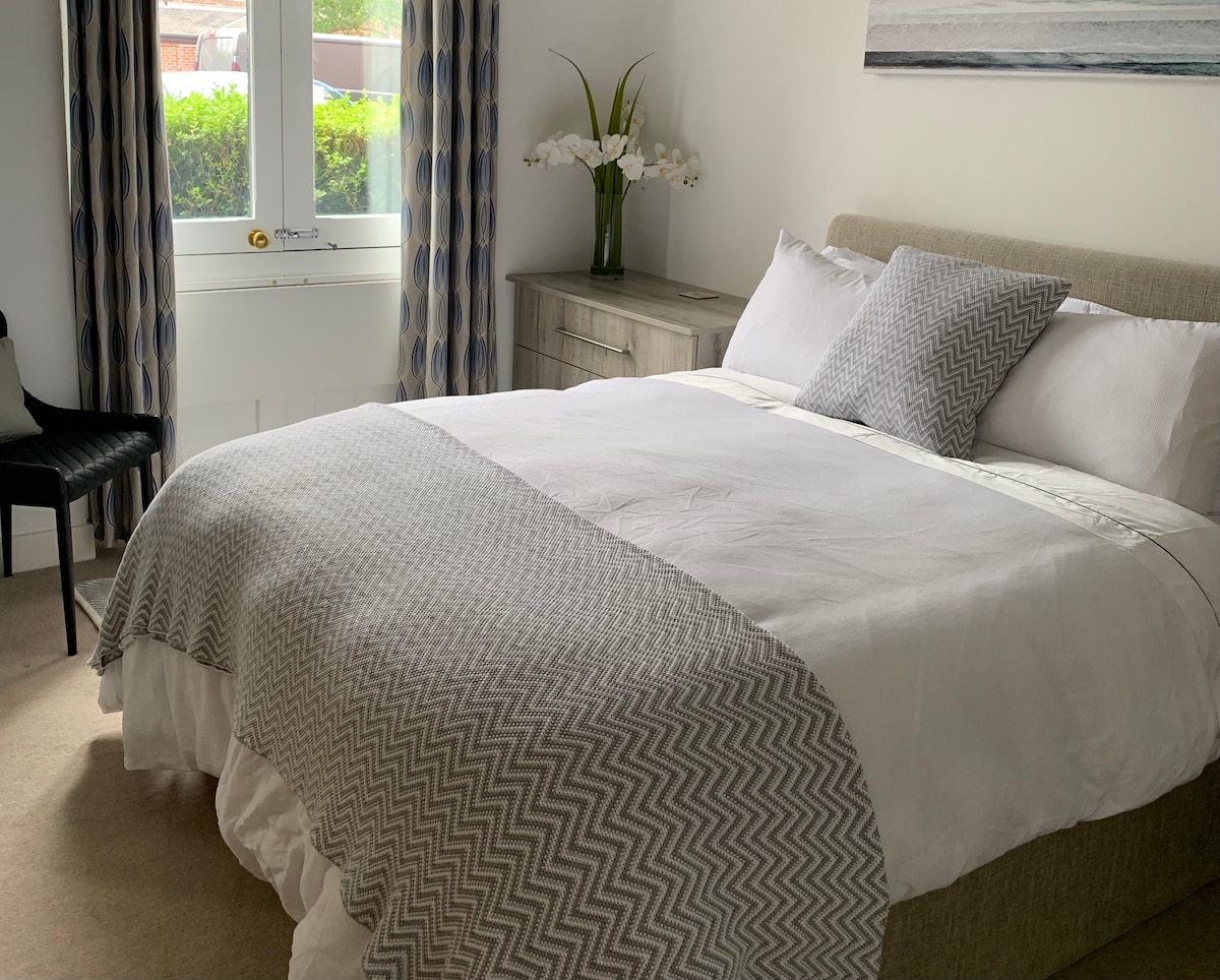 A cozy bedroom features a spacious bed adorned with a light grey chevron-patterned blanket. Soft lighting illuminates the room, complemented by stylish curtains and a decorative plant atop a simple dresser.