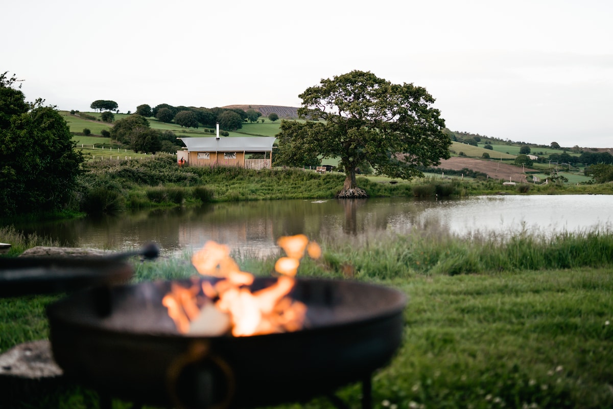 A fire pit with glowing flames is placed in the foreground, overlooking a tranquil pond. In the background, a wooden safari tent is set against the rolling countryside and a tree, adding natural beauty to the peaceful setting.