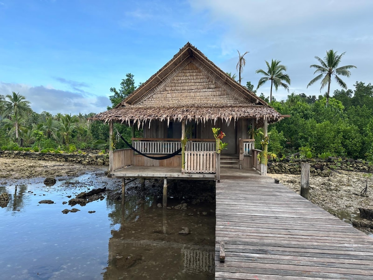 A water bungalow crafted from local wood features a thatched roof and a welcoming wooden terrace. A private pier extends over calm waters, surrounded by lush greenery and palm trees. The serene scene is complemented by a clear blue sky.