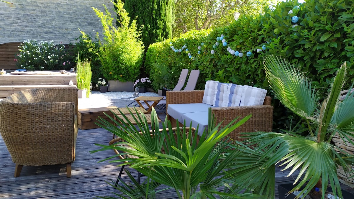 A serene outdoor space features a comfortable seating area with woven furniture, including a spacious sofa and armchairs. Lush greenery surrounds the area, complemented by decorative lights strung above. A wooden deck provides a relaxing atmosphere, highlighted by potted plants and a cozy ambiance.