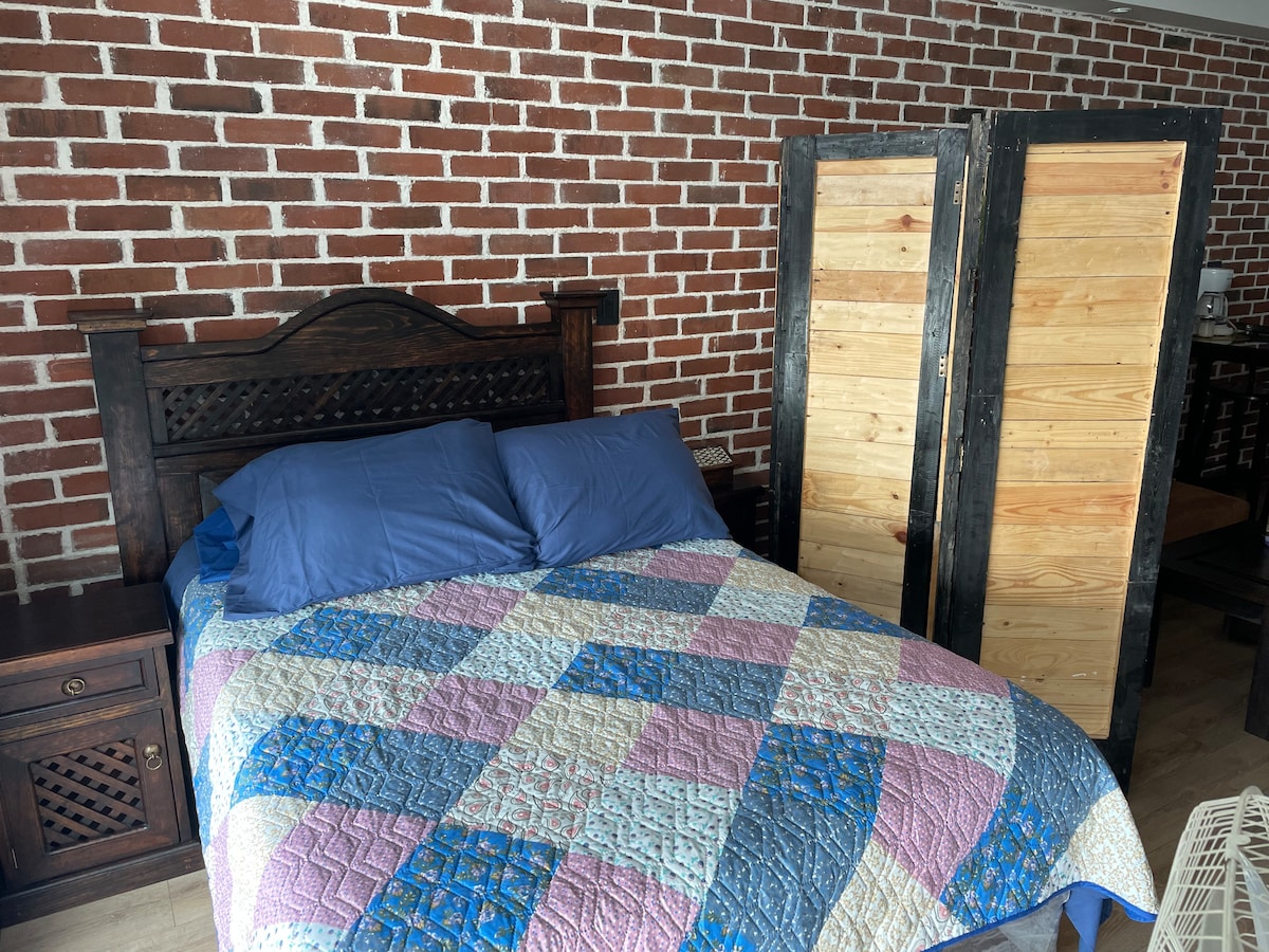 A spacious bed is positioned against a textured brick wall, covered with a patchwork blanket in shades of blue, pink, and cream. Two wooden screens stand nearby, adding privacy to the sleeping area.