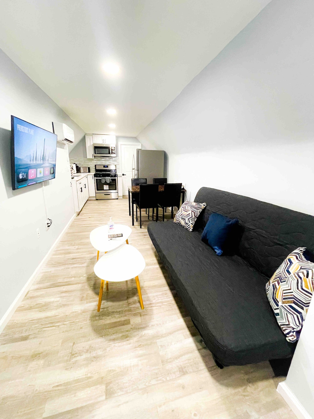 The living area features a dark sofa that can convert into a double bed, accented by decorative pillows. A small coffee table, along with a dining table for four, is positioned near a modern kitchen equipped with stainless steel appliances and overhead lighting.