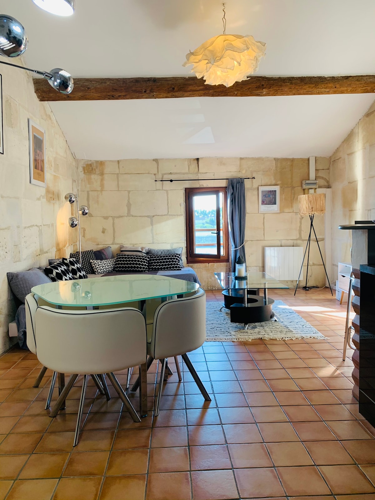 A living area features a mix of natural stone walls and modern furnishings. A round glass dining table is surrounded by white chairs, while a soft rug lies beneath. A large window offers views of the river, allowing natural light to fill the space.
