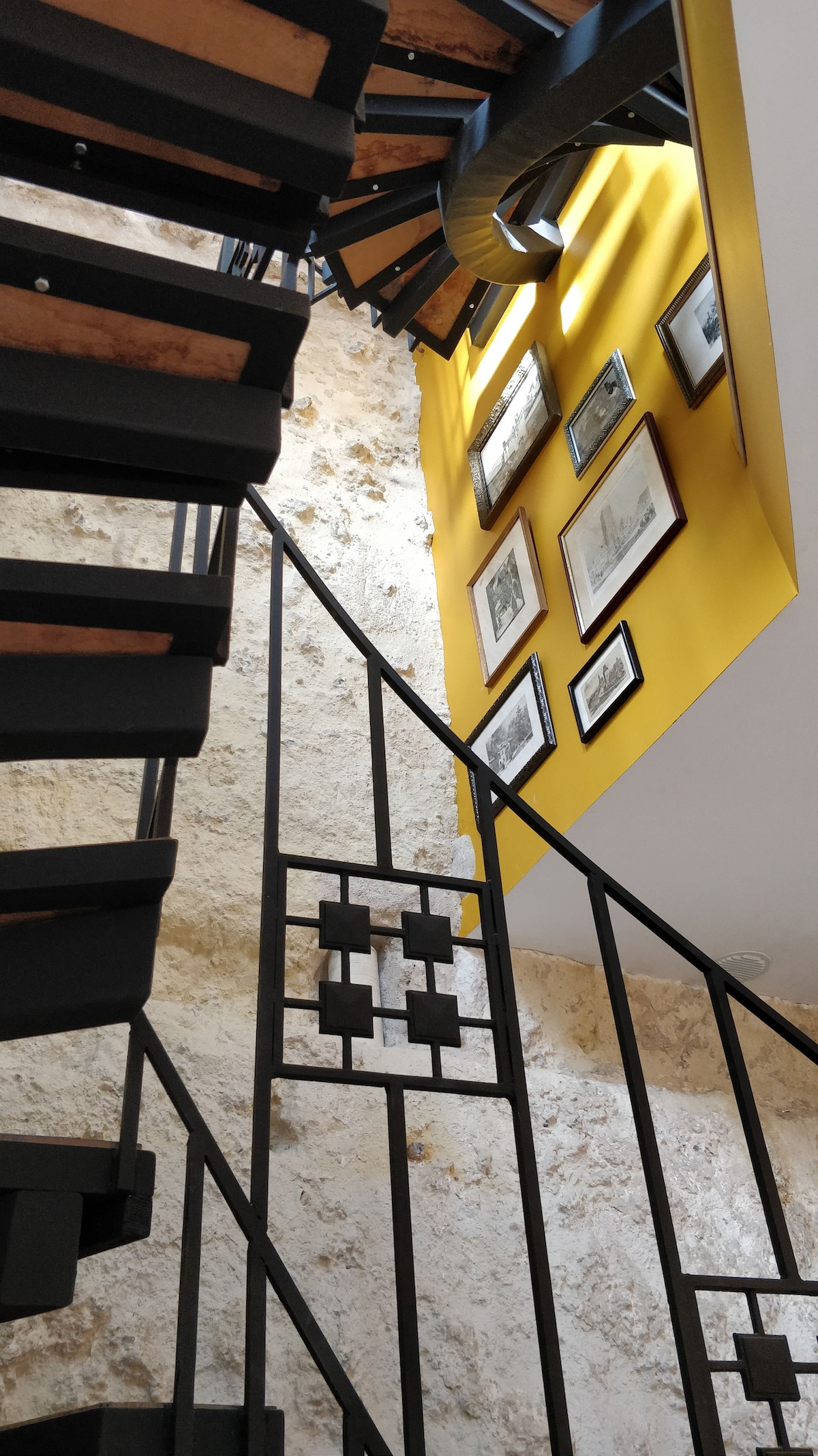 A spiral staircase features a metal railing with a geometric design, leading to a brightly lit area above. The wall is painted in yellow and adorned with several framed black-and-white photographs, enhancing the unique character of the interior.