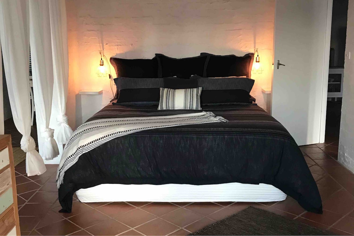 A spacious sleeping area features a large bed dressed in dark linens and accented with decorative pillows. Soft lighting is provided by pendant lights mounted on either side of the bed. A throw blanket is elegantly draped across the foot of the bed, enhancing the inviting atmosphere.
