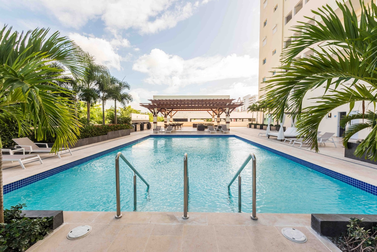 A serene swimming pool is framed by lush palm trees, with stainless steel handrails leading into the water. A shaded pergola offers seating and dining areas nearby, creating a relaxing space. The clear blue sky is reflected on the pool's surface, enhancing the tranquil ambiance.