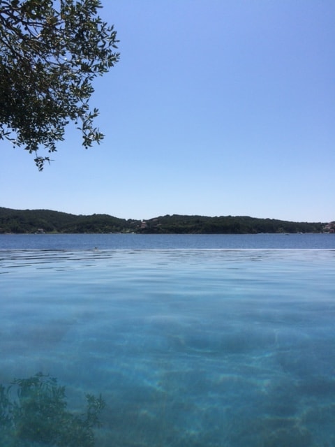 The image captures a serene view of the calm sea, with the clear water blending into the horizon. Lush green hills are visible in the distance under a clear blue sky, creating a peaceful atmosphere. The foreground features the tranquil surface of the infinity pool reflecting the vibrant surroundings.
