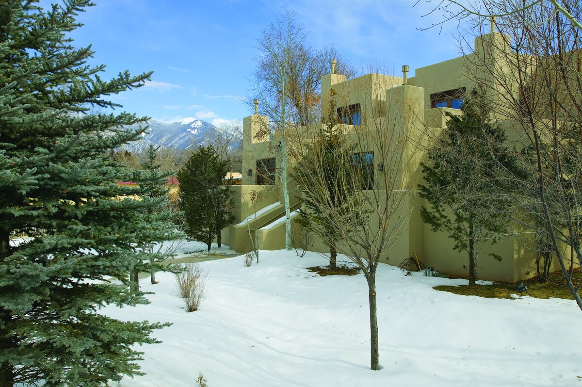 A multi-story building is set against a backdrop of snow-covered ground, with trees partially bare and a clear blue sky. The architectural design features soft, earthy tones and geometric shapes, harmonizing with the natural landscape and distant mountains.