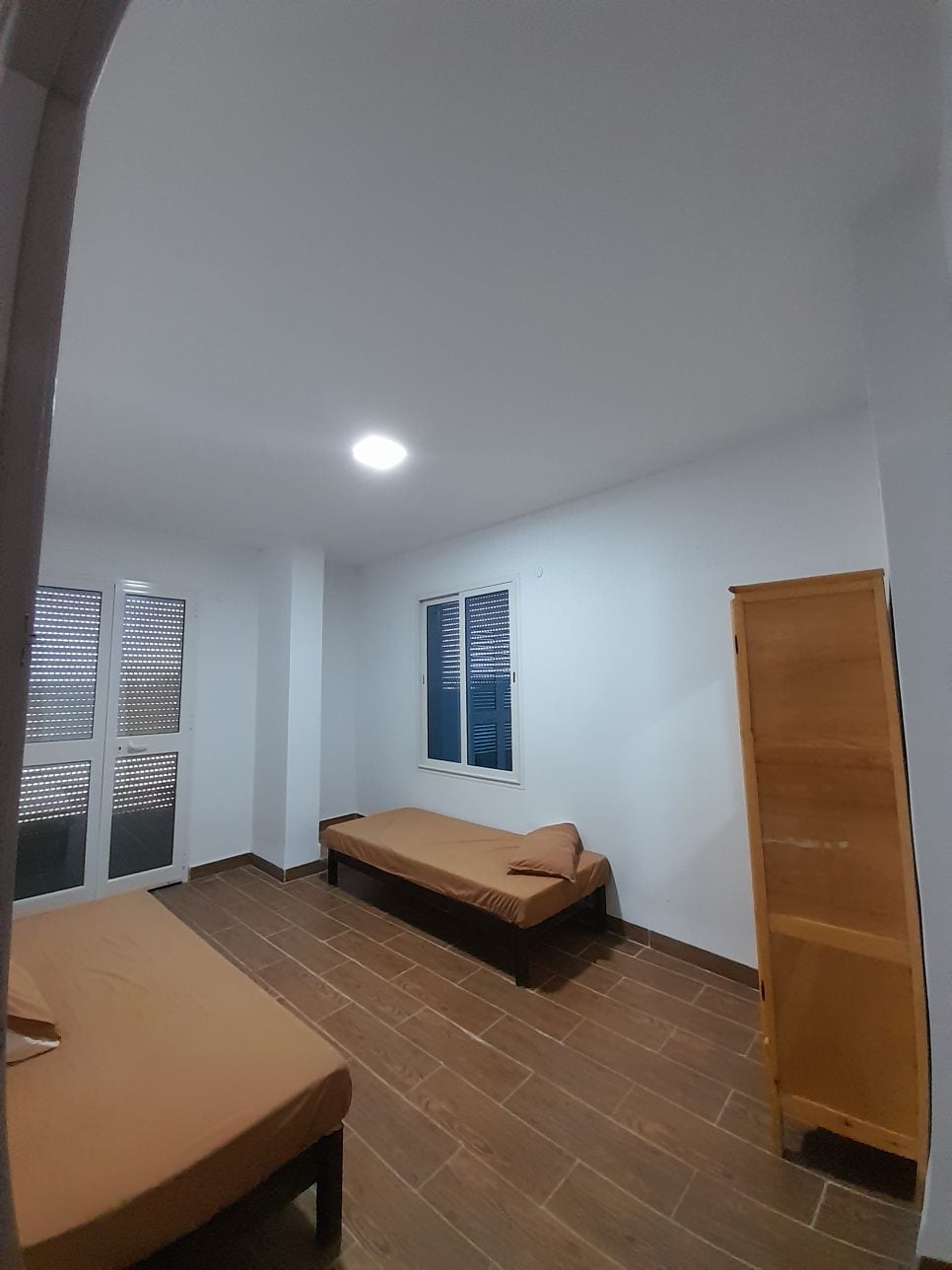 A well-lit room features two single beds with neutral bedding, positioned beside a window with wooden shutters. A wooden shelf stands against the wall, providing storage options. The smooth, tiled floor complements the clean, minimalist design of the space.