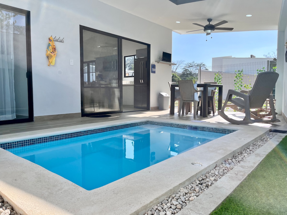 A private outdoor area features a clear blue pool complemented by a shaded seating area. Light wood furniture surrounds the space, and a ceiling fan is visible above. Glass doors provide a view of the interior, creating a seamless transition between indoor and outdoor living.