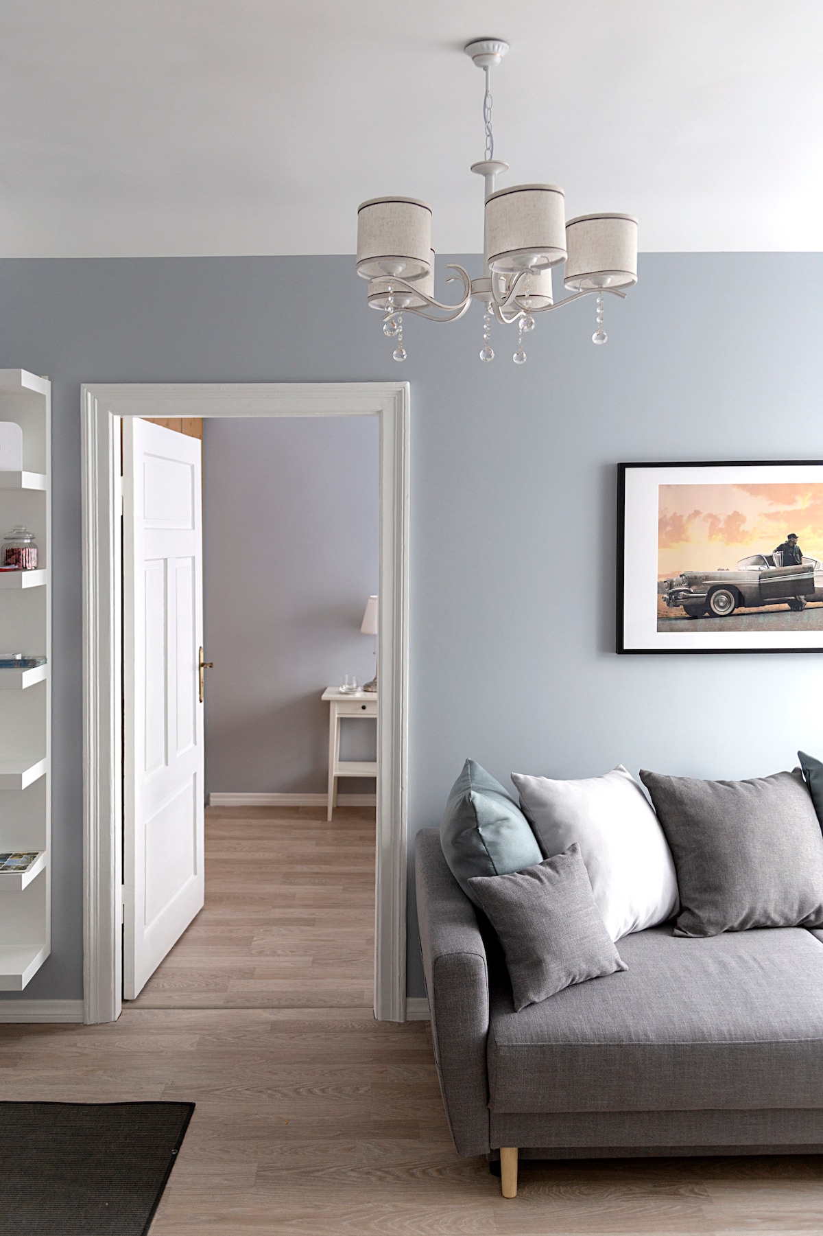 A cozy living area contains a modern gray sofa adorned with white and blue cushions. A delicate chandelier hangs from the ceiling, illuminating the space. An open door leads to a softly painted room, while a framed artwork adds a touch of character to the wall.