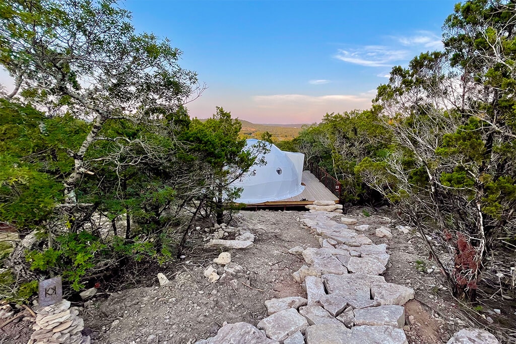 A winding stone pathway leads to a geodesic dome nestled among lush greenery, surrounded by tall shrubs and trees. The dome's white exterior contrasts with the natural landscape, creating a serene atmosphere. The distant horizon is visible under a soft blue sky.