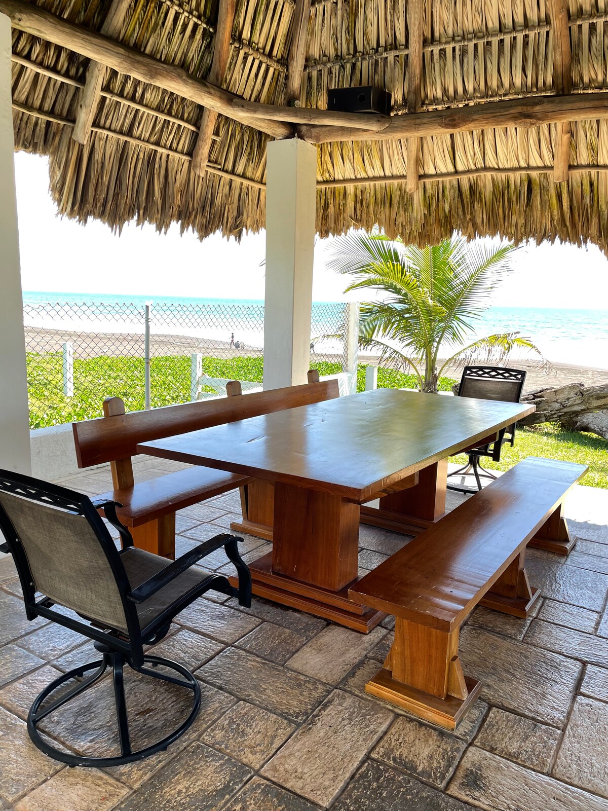 A shaded outdoor dining area features a wooden table with attached benches, complemented by a separate black metal chair. The space is set beneath a thatched roof, allowing views of the beach and ocean in the background, surrounded by a fenced garden.