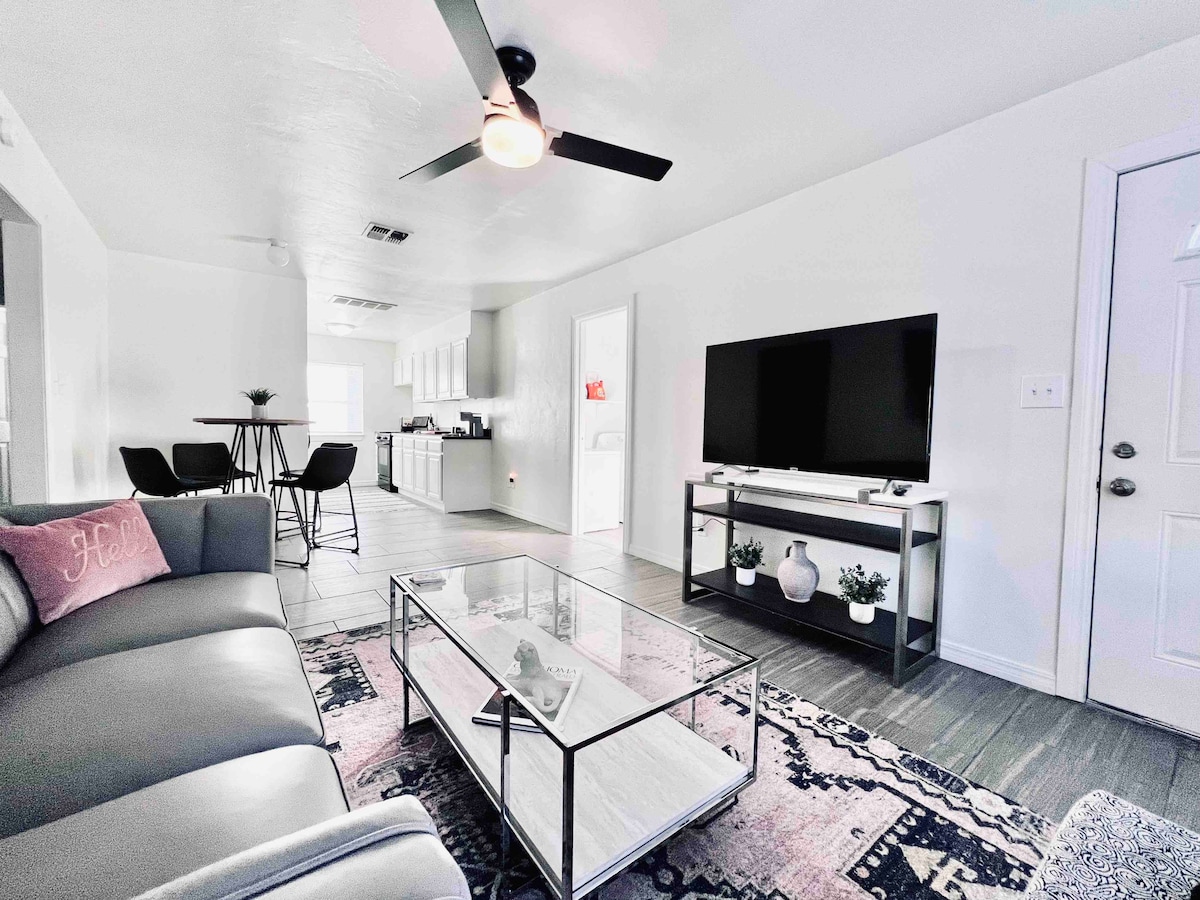 A modern living space features a comfortable gray sofa and a glass coffee table placed on a patterned area rug. A flat-screen TV is mounted on the wall, and stylish bar stools accompany a small dining table in the background, enhancing the open layout.