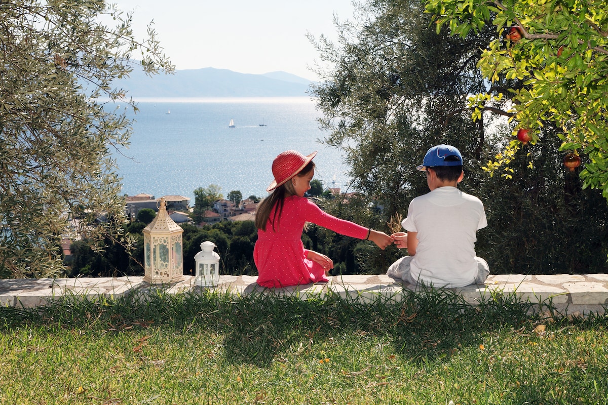 Two children sit on a stone wall overlooking the sea and distant islands. The sun reflects off the water, creating a shimmering effect. Olive trees and colorful flowers frame the scene, with a lantern placed on the grass nearby, hinting at a relaxed outdoor setting.