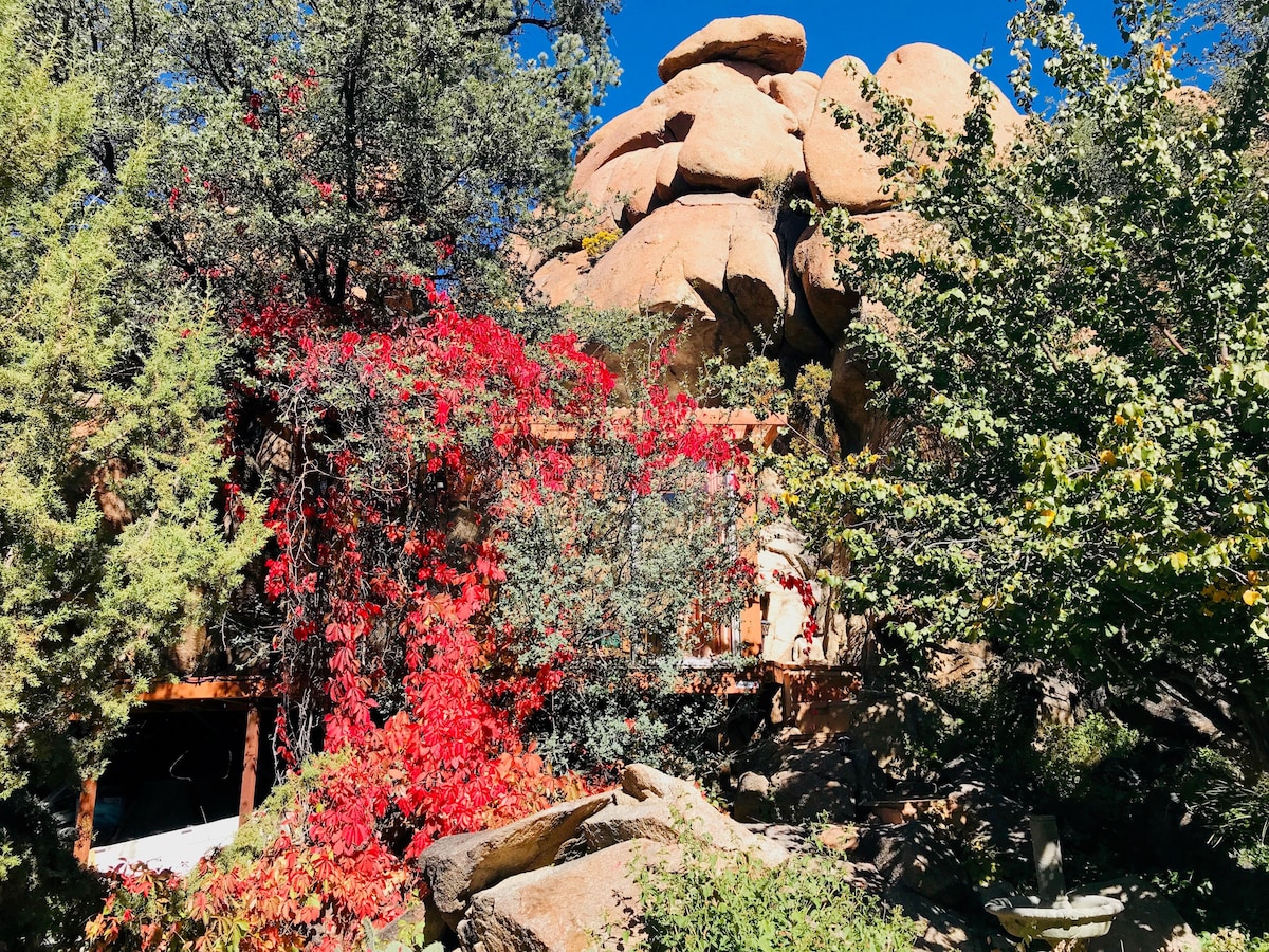 The exterior of the Treehouse is surrounded by vibrant foliage and rugged boulders. Red and green plants create a natural frame, complementing the earthy tones of the rocks and the structure. Clear blue skies provide a bright backdrop to this tranquil retreat.