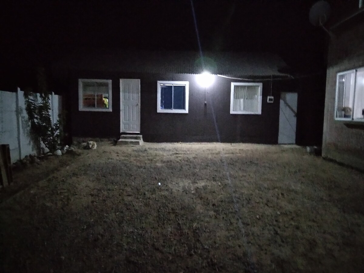 A nighttime view of the cabin exterior is presented, featuring a well-lit entrance with a white door and two windows. The surrounding area is characterized by gravel and sparse vegetation, illuminated by a bright overhead light. A fence encloses the property, enhancing privacy.