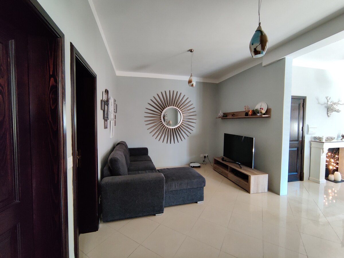 A living area featuring a gray sectional sofa is complemented by a round decorative mirror on the wall. Natural light enhances the spaciousness, while a television sits atop a light wooden console. The floor is tiled and the walls are painted in soft gray tones.