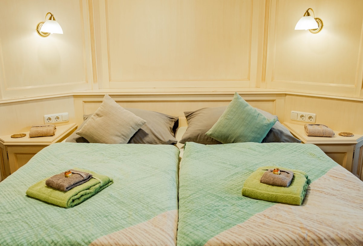 A spacious bed is dressed in light green and beige bedding, with two pillows positioned against a patterned headboard. Neatly folded towels are placed at the foot of the bed, complementing the color scheme. Soft lighting from wall-mounted lamps enhances the overall ambiance.