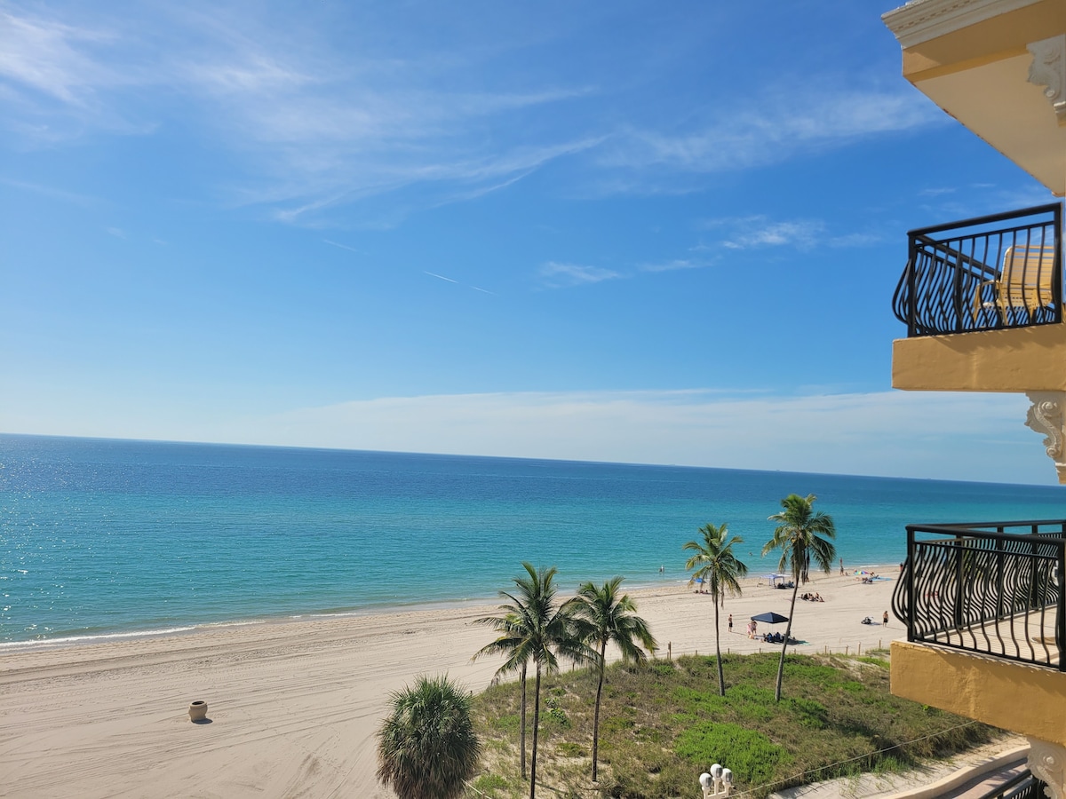 A clear view of the oceanfront showcases a sandy beach bordered by gentle waves. The sky is bright blue, complementing the turquoise water. A few palm trees are visible, providing a tropical feel, while beach umbrellas can be seen in the distance, indicating a relaxed beach atmosphere.