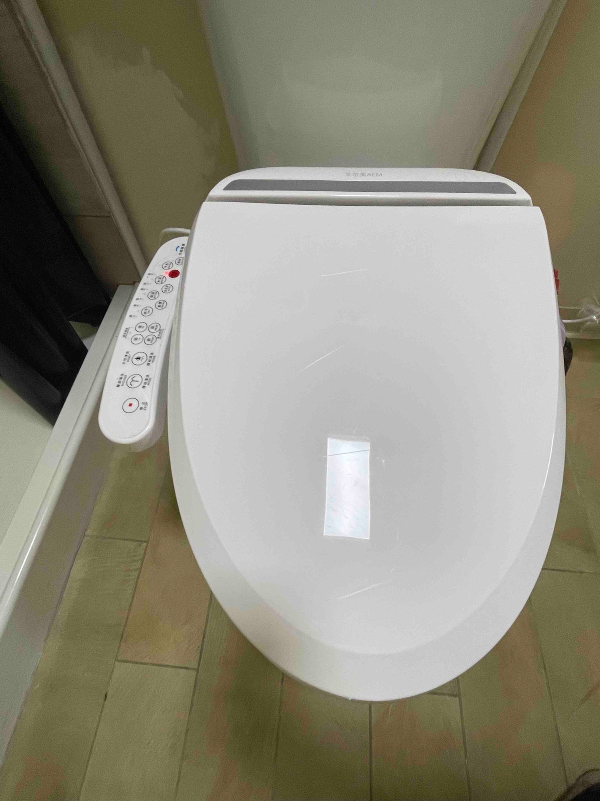 A modern toilet features a sleek design and is equipped with a remote control on the side, displaying various options. The surrounding area includes neutral-colored flooring, adding to the contemporary aesthetic of the bathroom space.