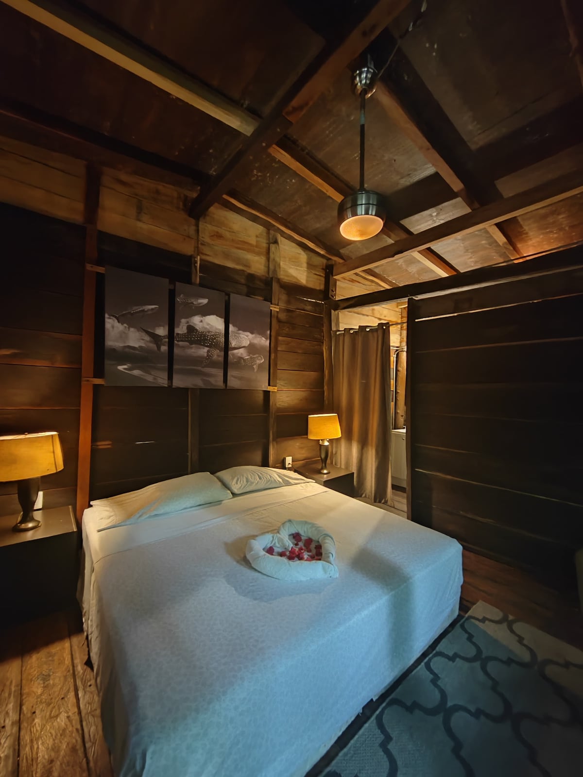 A cozy bedroom features a spacious bed topped with white linens and adorned with a decorative arrangement of flowers. Two bedside lamps provide soft lighting against the dark wooden walls. A curtain separates the sleeping area from the adjoining space, adding privacy.