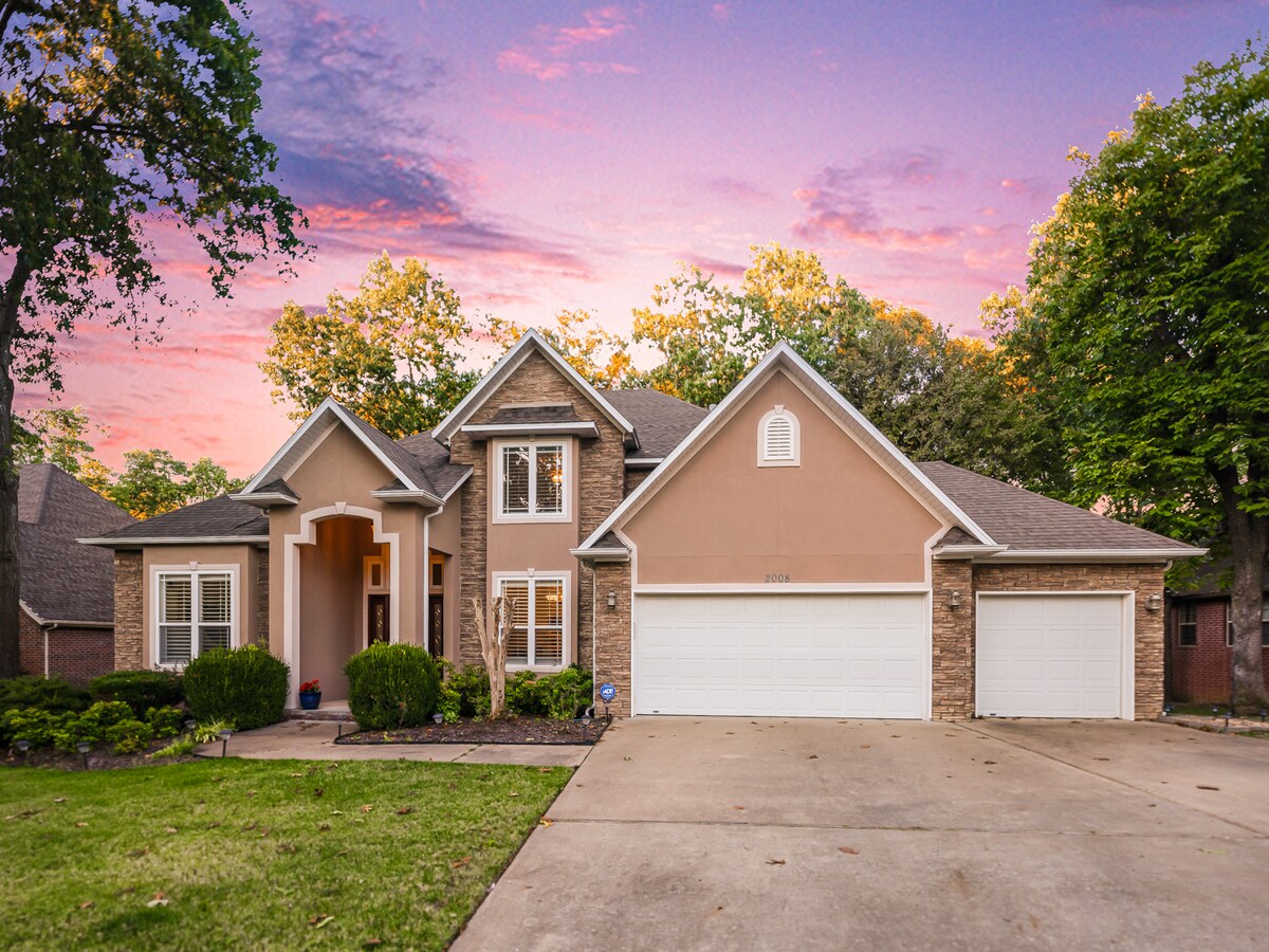 A single-story cottage exterior showcases a blend of warm-toned siding and natural stone accents. Lush greenery surrounds the entrance, which features a covered porch and large windows. The driveway leads to a spacious two-car garage, with trees providing a backdrop under a colorful sky.