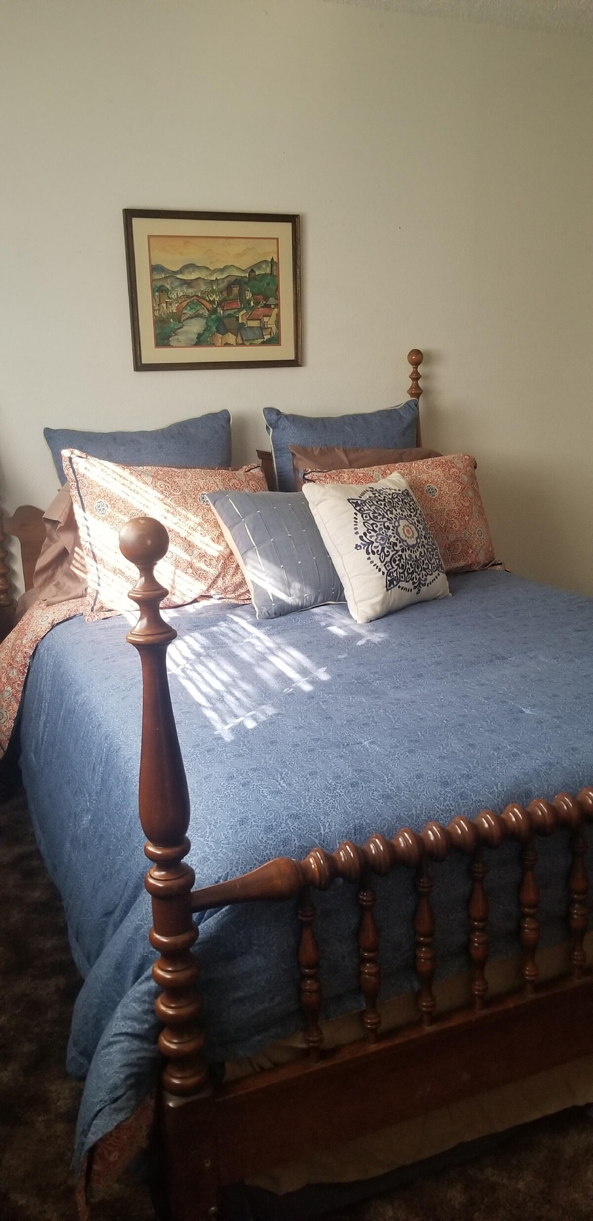 A cozy sleeping area features a wooden bed frame with a blue coverlet. Decorative pillows add texture against a backdrop of light-colored walls. A framed artwork hangs above, and natural light filters through a nearby window.