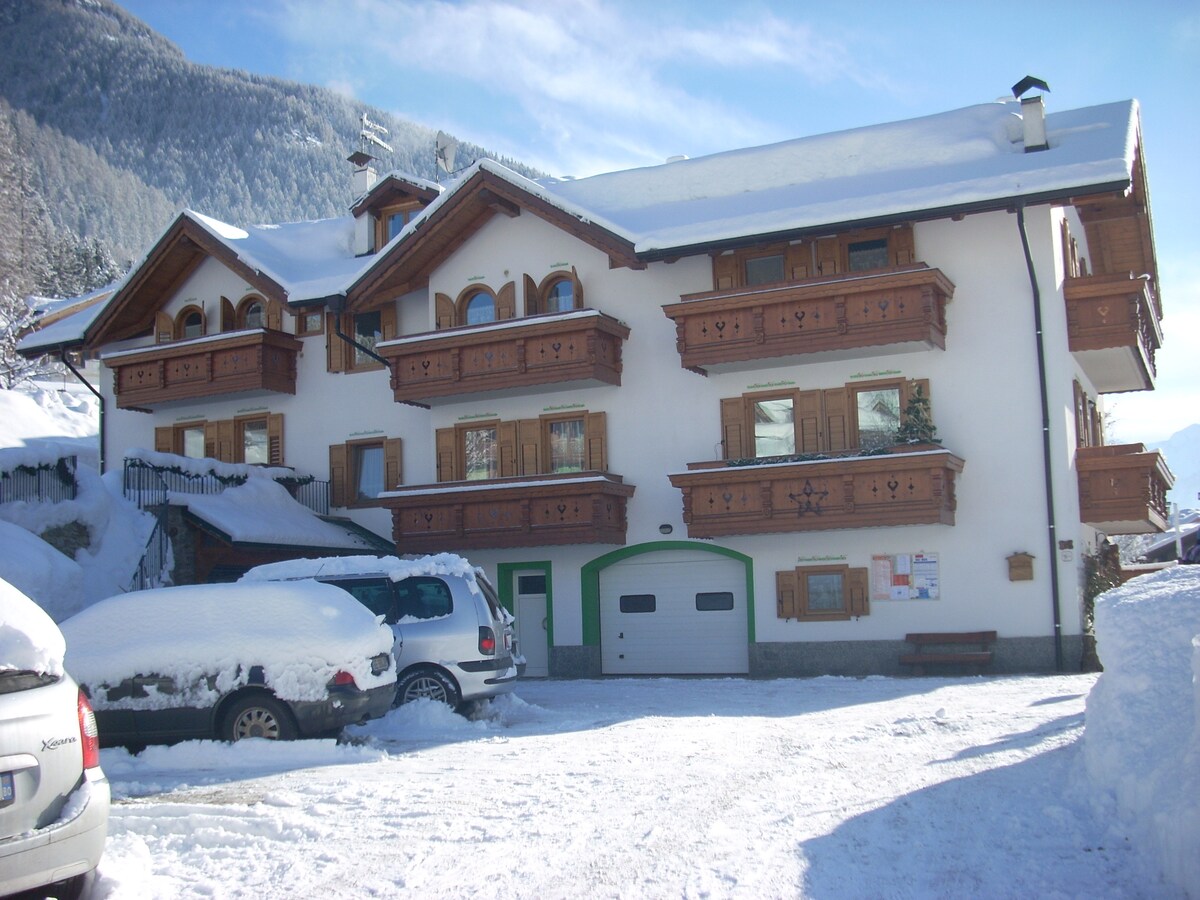 The exterior of a mountainous building is covered in snow, showcasing wooden balconies and multiple arched windows. A parking area with several vehicles is visible, along with a garage door. The surrounding landscape features snow-capped mountains under a clear blue sky.
