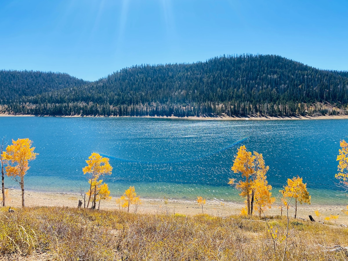 A serene lake is surrounded by trees with vibrant yellow foliage, set against a backdrop of rolling green hills. The clear blue water reflects sunlight, creating shimmering patterns on the surface. The sandy shoreline is visible in the foreground, enhancing the tranquil atmosphere.