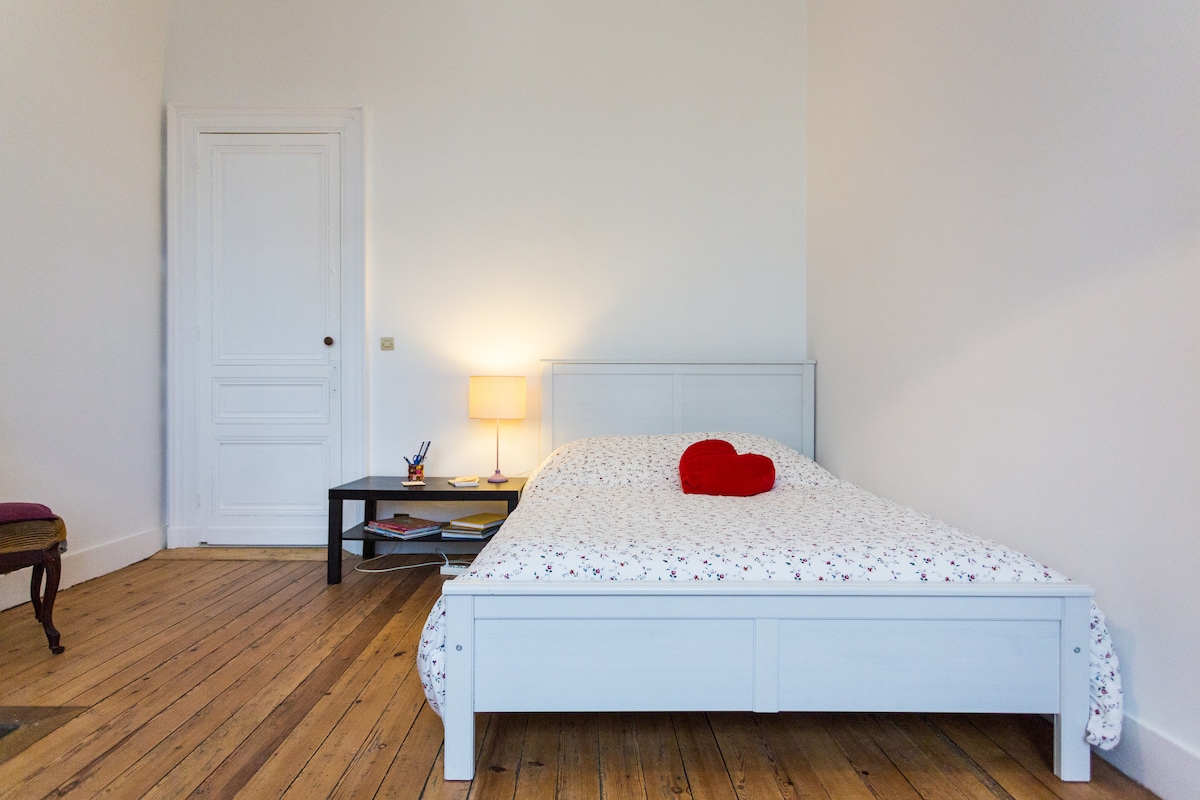 A well-lit sleeping area features a double bed dressed in floral bedding, complemented by a heart-shaped decorative pillow. A small bedside table with a lamp provides soft illumination, and a desk can be seen against the wall, offering a quiet space for work or reflection.