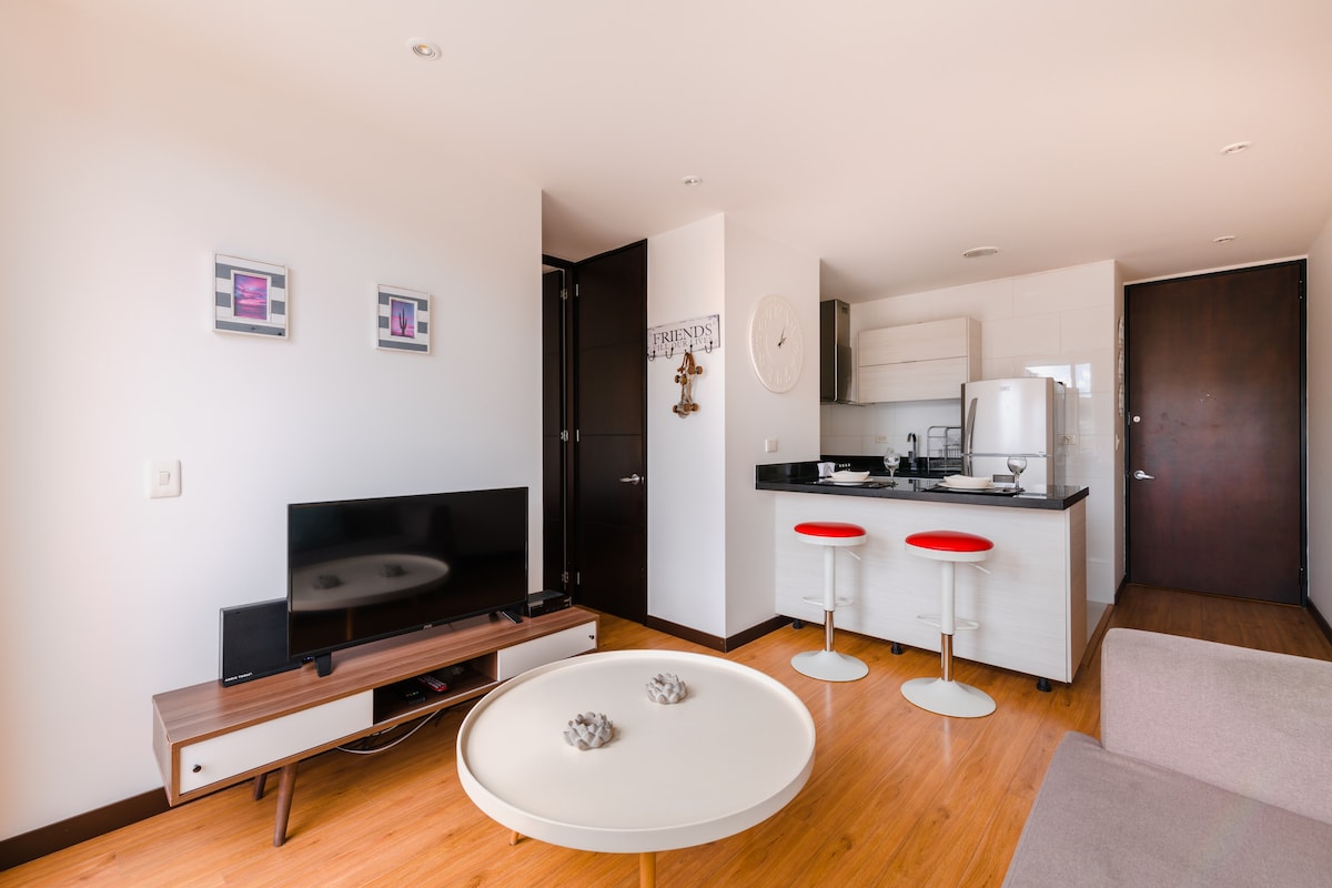 A modern living space is presented with a sleek television on a light wood media console. Red bar stools are positioned at a black-topped kitchen island, which complements the open layout. Light-colored walls and wood flooring create a bright and airy atmosphere.