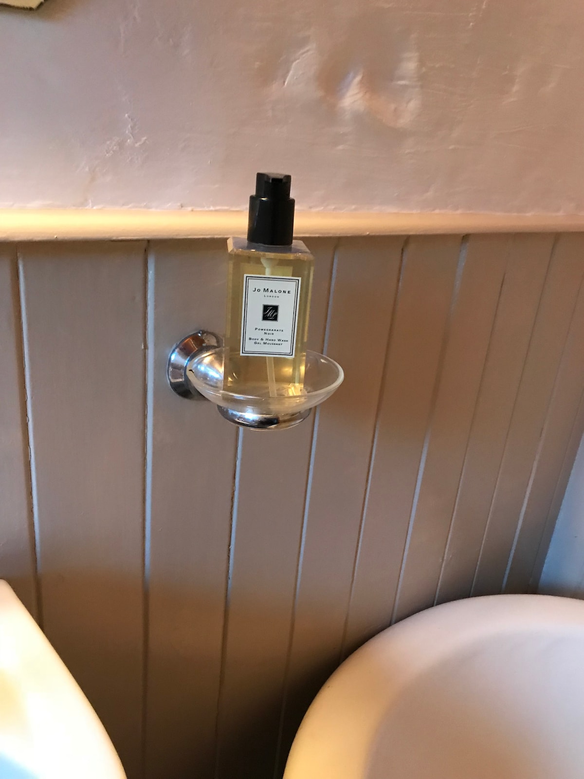 A sleek fragrance bottle is displayed on a glass holder attached to the pale wooden wall. The setting features pale wall paneling and simple, minimalist fixtures, providing a clean and tidy appearance. The surface beneath the holder is part of the bathroom's modern design.