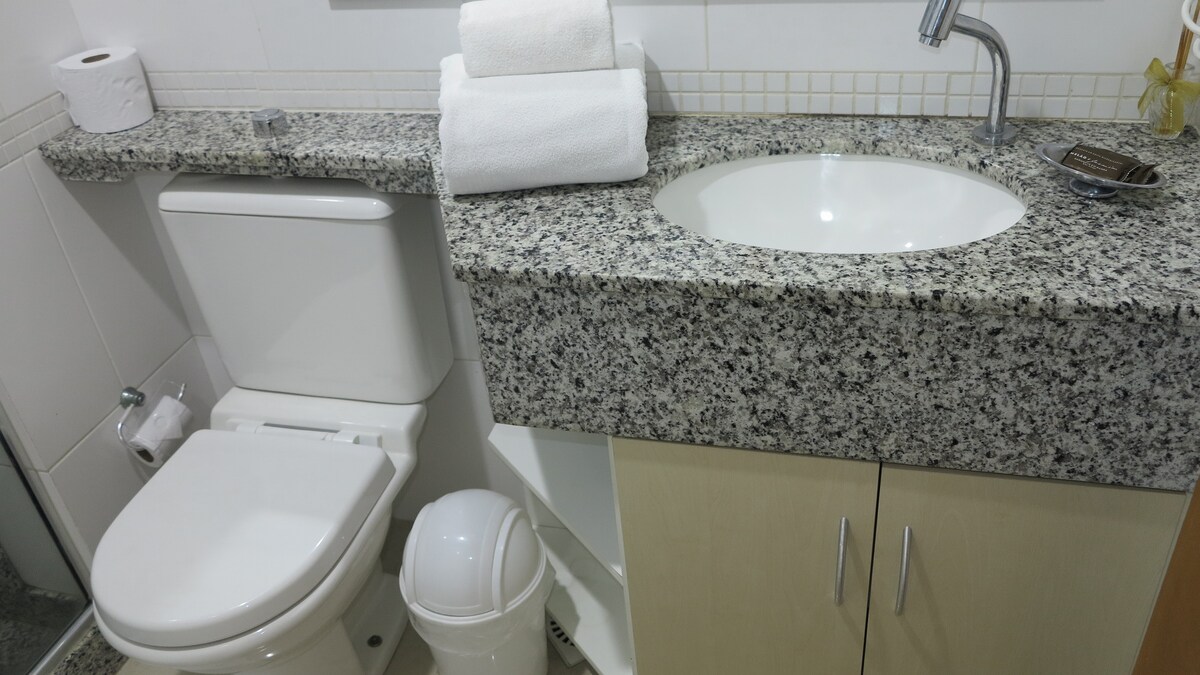 A compact bathroom is features a modern sink with a smooth granite countertop, complemented by rolled white towels. A toilet is positioned adjacent to a cabinet, providing storage space below. Accessories include a toilet paper roll and a waste bin for convenience.